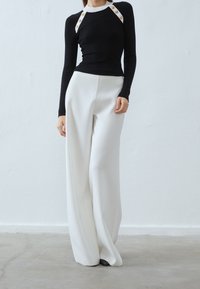 Haut à manches longues noir côtelé avec col blanc et accents de boutons dorés, associé à un pantalon large blanc en tissu lisse. Design simple et élégant.