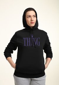 Felpa nera con cappuccio caratterizzata da un testo grafico viola "THING" sulla parte anteriore. Tessuto liscio con polsini e orlo a costine, indossata con pantaloni della tuta grigi.