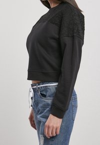 Femme portant un sweat-shirt court noir à manches longues avec des détails en dentelle sur les épaules, associé à un jean en denim bleu et une ceinture blanche à cordon.