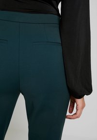 Teal passande byxor med en slät textur, med två bakfickor. En svart långärmad topp är delvis synlig.