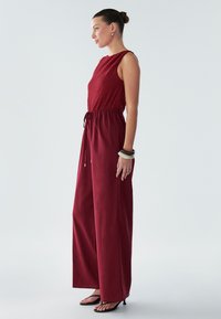 Bordo kombinezon z teksturirano zgornjim delom in elastičnim pasom, z razširjenimi hlačnicami in sproščenim krojem. Nošen z enostavnimi črnimi sandali.