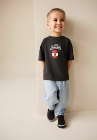 Schwarzes T-Shirt mit Spider-Man-Grafik, hellblaue weite Jeans und schwarze Sneakers. Steht lächelnd vor einer cremefarbenen Wand.