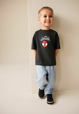 Fekete póló Pókember grafikával, világoskék bőszárú farmer, és fekete sportcipő. Krémszínű fal előtt áll, mosolyogva.