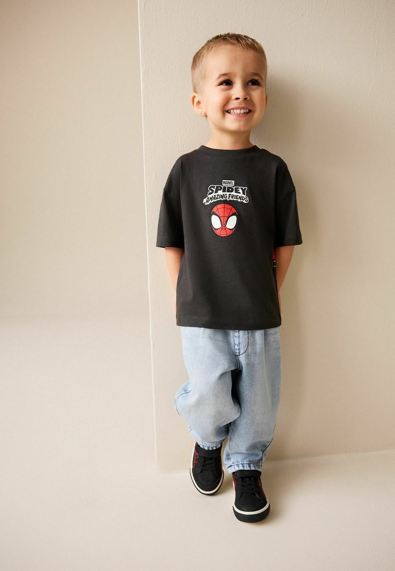 Schwarzes T-Shirt mit Spider-Man-Grafik, hellblaue weite Jeans und schwarze Sneakers. Steht lächelnd vor einer cremefarbenen Wand.