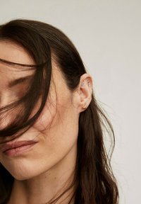 Pendientes de oro y plata con diseños variados, incluyendo una flor y una ala, lucidos en una oreja parcialmente oculta con cabello castaño suelto.