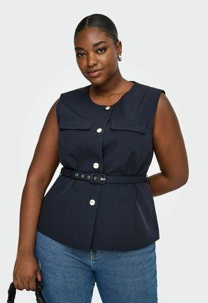 Žena s tmavou pokožkou a vlasmi stiahnutými dozadu, má na sebe tmavomodrú bezrukávovú vestu na gombíky s opaskom, modré rifle a drží čiernu kabelku.