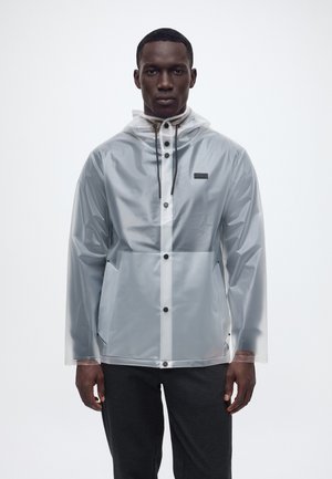 Bărbat purtând o jachetă de ploaie gri translucidă cu nasturi negri și șnururi de ajustare, stând pe un fundal alb simplu.