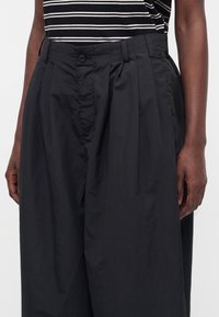 Personne portant un pantalon noir plissé ample et un haut sans manches rayé noir et blanc sur un fond clair uni.
