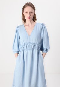 Rochie din chambray bleu deschis, cu decolteu în V, talie elastică, mâneci bufante și buzunare laterale. Textură netedă cu detalii simple de cusătură.