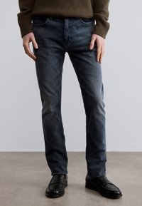 Mørkeblå denimjeans med smal passform, med subtil falming og fem-lomme-design, brukt med svarte skinnsko, kombinert med en brun genser.