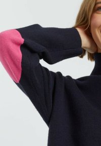 Pull marin avec un col haut et des coudières en rose. Matériel en laine texturé avec une coupe décontractée. Détails de couture visibles.