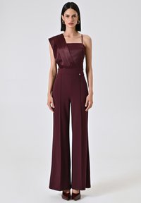 Combinaison bourgogne avec une bretelle en satin, décolleté carré et silhouette évasée ; présente des plis sur les côtés et un bouton décoratif.
