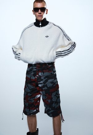 Modell kannab valget karvast Adidas lühikest tõmblukuga jopet mustade triipudega ja kamuflaažvärvilisi lahtisi pükse, seisab käed puusal.