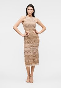 Femme debout, mains sur les hanches, portant une robe midi beige et marron sans manches à motif ondulé et des talons ouverts dorés.