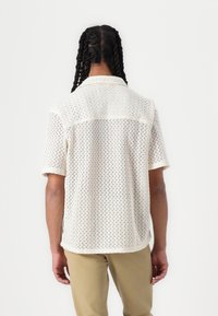 Chemise crème à manches courtes avec un motif tricoté en zigzag, dotée d'un col et d'un empiècement au dos. Portée avec un pantalon beige clair.