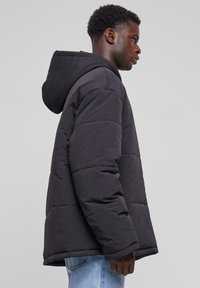 Sort polstret jakke med hætte, der har quiltede sektioner og lange ærmer. Materialet ser blødt og glat ud, båret over blå jeans.