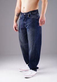 Dark blue denim jeans with a straight cut, five pockets, and metal button accents. Worn with white sneakers.