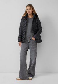 Zwarte pufferjack met capuchon, voorzien van een gewatteerd patroon. Draagt over een grijze sweater en gecombineerd met wijde grijze jeans en lichte sneakers.