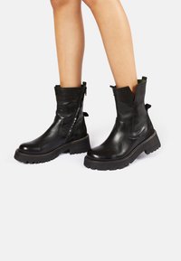 Botas de tobillo de cuero negro con una suela gruesa baja, cremallera lateral y diseño cortado en la parte superior. Cuenta con un acabado texturizado.