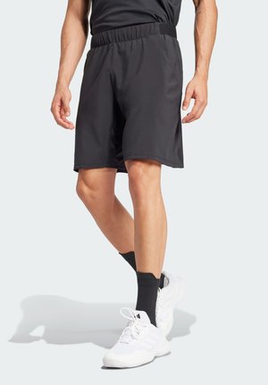Calções atléticos pretos com uma cintura elástica e bolsos laterais, feitos de um material leve, apresentados com meias pretas e sapatos brancos.