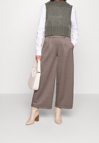 Olivgrüner, kurz geschnittener Pullover über einem weißen Hemd, kombiniert mit karierter, weit geschnittener Hose. Das Model trägt eine cremefarbene Handtasche und beige Knöchelstiefel.