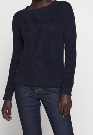 Personne à la peau foncée portant un pull en tricot côtelé bleu marine à manches longues et un jean bleu foncé, debout devant un fond clair.