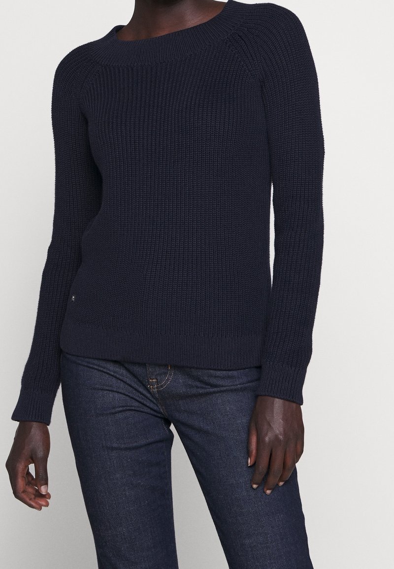 Personne à la peau foncée portant un pull en tricot côtelé bleu marine à manches longues et un jean bleu foncé, debout devant un fond clair.
