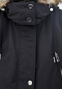 Czarny zimowy płaszcz z kapturem wyściełanym futrem, wyposażony w dwa duże guziki, zamek błyskawiczny oraz sznureczki przy kołnierzu umożliwiające regulację dopasowania.