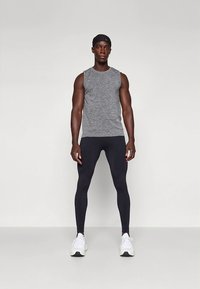 Sportoutfit med en grå ärmlös topp och svarta leggings. Modellen bär vita sneakers och ett svart pannband, stående mot en neutral bakgrund.