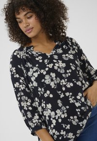 Blouse florale avec un fond marine et des motifs floraux blancs, décolleté en V et manches trois-quarts en un tissu léger et texturé.