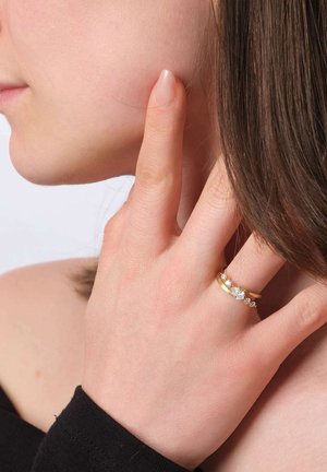 Gold ring with three sparkling gemstones, including a larger center stone, worn on a finger near a face. Smooth surface, delicate design.