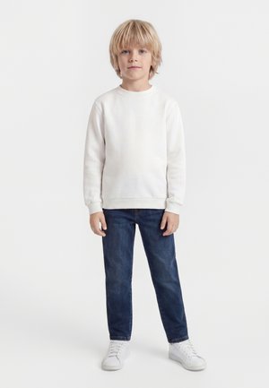 Lyshåret dreng står vendt fremad iført en hvid langærmet skjorte, blå jeans og hvide sneakers foran en ensfarvet hvid baggrund.