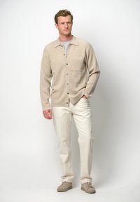 Beige gestrickte Strickjacke mit einer Tasche und Knöpfen, kombiniert mit weißen Hosen und hellgrauen Schuhen, vor einem schlichten Hintergrund stehend.