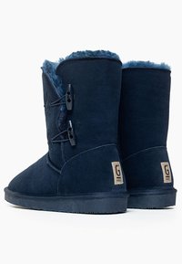 Botas de ante azul marino con un suave forro de piel de oveja, de altura hasta la mitad de la pantorrilla, con toggles decorativos y un parche con el logotipo en el lateral.