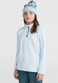 Pull-over en polaire bleu clair avec un col zippé à moitié, orné d'un logo bleu marine. Porté avec un bonnet bleu assorti avec un pompon.