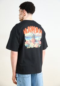 Černé tričko s krátkým rukávem s barevným grafikou "Hawaii Aloha", ananasem, květinami a horami na zadní straně.