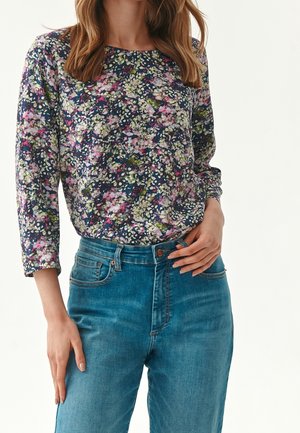 Kvinde iført en langærmet bluse med blomsterprint, stukket ned i højttaljede blå jeans, stående med den ene hånd på hoften.