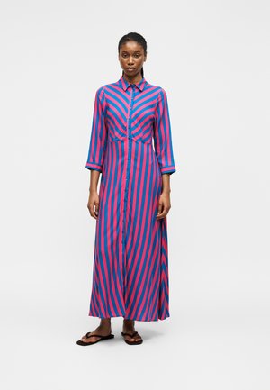 Femme portant une robe boutonnée à manches longues, longueur cheville, rayée bleu et rose, avec des sandales noires, debout devant un fond uni.