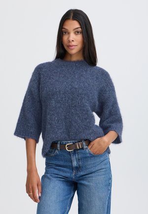 Femme aux longs cheveux foncés portant un pull bleu duveteux et un jean bleu avec une ceinture noire, debout devant un fond clair et uni.