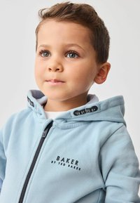 Šviesiai mėlynas užtrauktuko džemperis su juodais akcentais. Savybės: "BAKER BY TED BAKER" logotipas ir minkšta tekstūra. Kapšas išklotas pilka medžiaga.