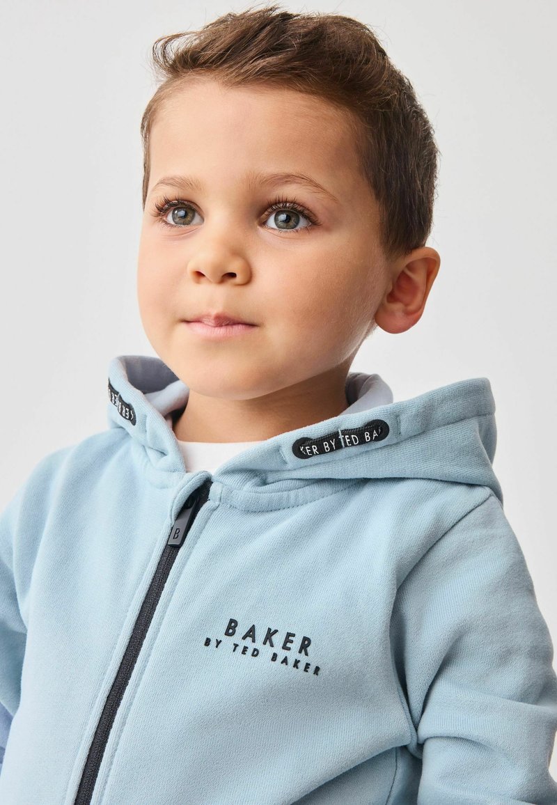 Šviesiai mėlynas užtrauktuko džemperis su juodais akcentais. Savybės: "BAKER BY TED BAKER" logotipas ir minkšta tekstūra. Kapšas išklotas pilka medžiaga.