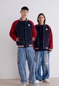 Marineblå og bordeaux varsityjakker med hvide stribede manchetter, forsynet med et broderet logo, sammen med løse denimbukser.