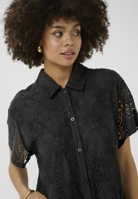 Black button-up shirt with lace overlay featuring floral patterns, short sleeves, and a structured collar. Textured and semi-sheer design.
