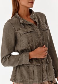 Olivgrön jacka med figursydd design, utrustad med två bröstfickor, metallknappar och skärp i midjan. Texturerat tyg med lätt utseende.