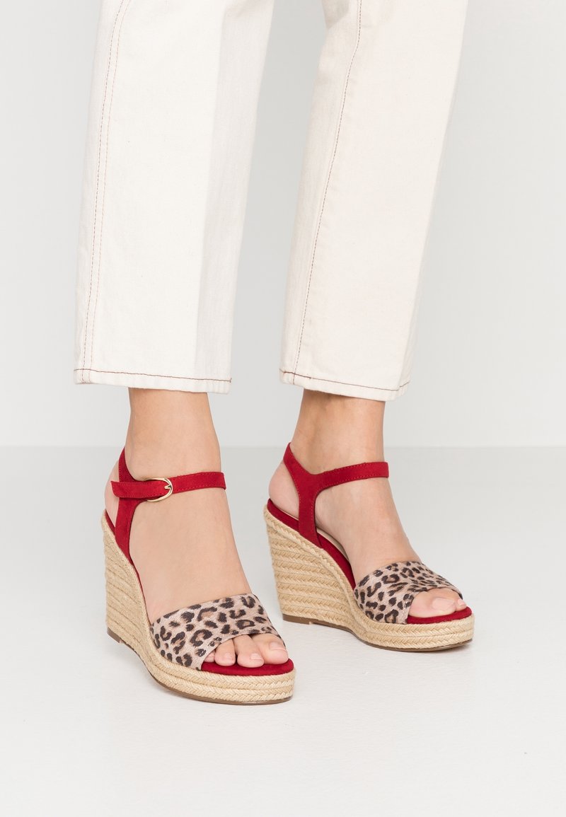 Pieds portant des sandales compensées rouges avec des brides imprimé léopard et des boucles à la cheville, associées à un pantalon court de couleur claire.