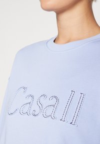 Sweatshirt bleu deschis cu guler rotund, având brodat textul "Casall" cu fir întunecat în contrast. Material moale, cu o croială lejeră.