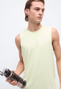 Jeune homme portant un débardeur jaune pâle de la marque Puma, tenant une bouteille d'eau transparente avec un couvercle noir, regardant vers sa droite.