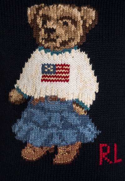 Pleteni pulover z motivom medveda v belem puloverju z ameriško zastavo, modro denim krilo in rjavimi škornji na temnem ozadju. Rdeč detajl "RL".