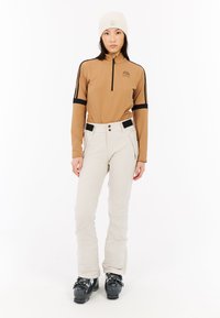 Długi rękaw z zamkiem w kolorze tan, z czarnymi akcentami, w połączeniu z beżowymi spodniami narciarskimi z guzikami i zamkami błyskawicznymi, w zestawie z czarnymi butami narciarskimi.