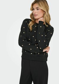 Zwarte sweatshirt met gouden hartpatroon, geribde boorden en zoom, ontspannen pasvorm. Gecombineerd met zwarte broek, gladde textuur.